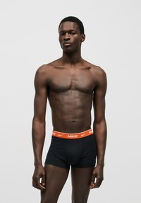 Αθλητικός άντρας στέκεται μπροστά από απλό φόντο, φοράει μαύρα εσώρουχα Nike με πορτοκαλί ζώνη στη μέση.
