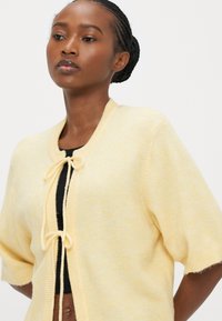 Light yellow cardigan with short sleeves, round neckline, and tie closure at the front. Soft, textured fabric with a subtle pattern.