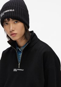 Sudadera negra con cremallera y cuello de canalé, con "KARL LAGERFELD JEANS" bordado en blanco. Llevada sobre una camisa de mezclilla azul. Gorro negro.