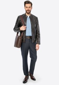 Uomo che indossa una giacca di pelle nera con dettagli a costine, un maglione blu, pantaloni scuri e scarpe marroni, mentre tiene una borsa a tracolla in pelle marrone.