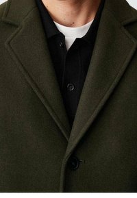 Donkergroen wollen jas met een gestructureerde kraag, enkele knoopsluiting en een textuurstof, gelaagd over een zwarte poloshirt.