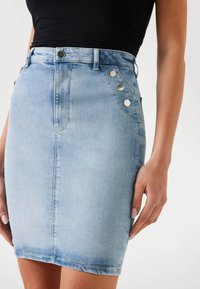 Helles blauer Jeansrock mit taillierter Passform, der eine glatte Textur, fünf Taschenstil und silberne Knopfakzente an der Seite aufweist.