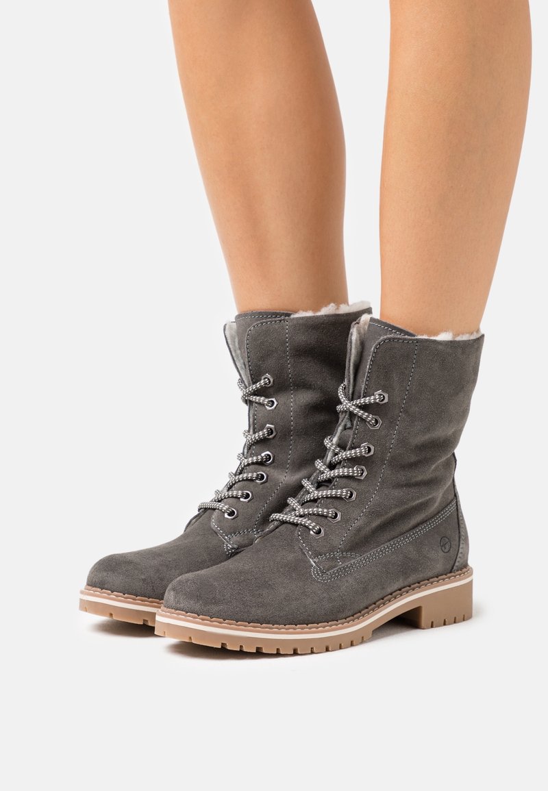 Bottes grises en daim à lacets avec semelles en caoutchouc beige et doublure polaire blanche, portées sur des jambes nues sur fond blanc.