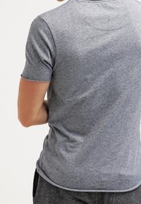 Osoba ubrana w gładką, szaro-fioletową koszulkę z krótkim rękawem, pokazana z tyłu z jedną ręką zgięta za ciałem.