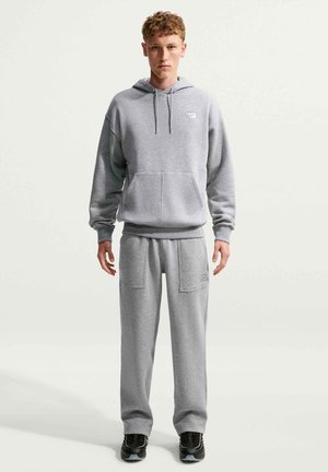 Giovane uomo in piedi che indossa una felpa con cappuccio grigia e pantaloni della tuta coordinati con sneakers nere su uno sfondo chiaro e semplice.