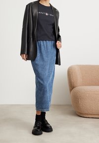 Person iført en sort læderjakke, sort logo-t-shirt, blå denim midi-nederdel, sorte sokker og sorte chunky loafers, der står indendørs.