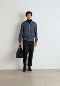 Grauer Reißverschluss-Cardigan, schwarzes Rollkragenoberteil, schwarze Hose, schwarze Schuhe und eine strukturierte schwarze Tasche. Glatter Stoff und schlankes Design.