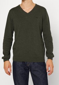 Mann trägt einen dunkelgrünen V-Ausschnitt-Pullover und eine dunkelblaue Jeans, steht vor einem einfarbigen Hintergrund mit entspannten Händen an den Seiten.