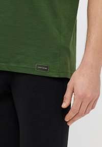 Πράσινο βαμβακερό t-shirt με στρογγυλεμένη κάτω ραφή, που διαθέτει μαύρη ετικέτα με το κείμενο "TOMMY HILFIGER" στην κάτω αριστερή γωνία. Ορατό μαύρο παντελόνι.