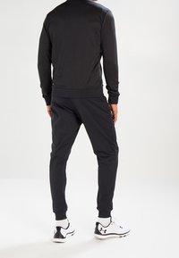 Schwarzer sportlicher Pullover und Hose, glattes Material, gerippte Bündchen, taillierte Passform. Tragen mit weißen und schwarzen Sportschuhen mit gepolsterten Sohlen.