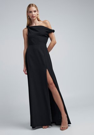 Robe noire asymétrique avec détail de nœud, fente sur le côté, longueur maxi, texture lisse et style minimaliste.