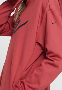 Sweat à capuche de sport rouge en tissu lisse, avec un grand logo noir sur le devant et un plus petit logo sur la manche. Capuche avec cordon de serrage.