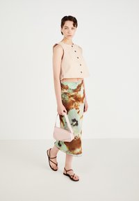 Top scurt de culoare roz deschis, cu nasturi negri, asortat cu o fustă lungă, fluidă, tie-dye în nuanțe de verde, maro și crem. Ținând în mână o geantă mică de culoare roz.