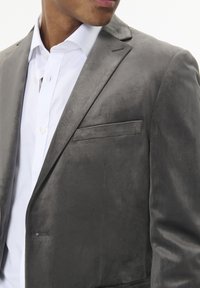 Pelēks zamšādas bleizeris ar vienu krūšu kabatu, ar izgriezumiem uz lapelēm un gludu tekstūru. Nēsāts pār baltu kreklu ar apkakli.