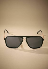 Schwarze rechteckige Sonnenbrille mit dunklen Gläsern, goldenen Metallakzenten an der Brücke und den Scharnieren sowie schwarzen matten Bügeln.