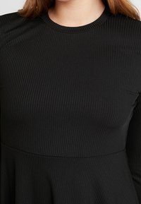 Musta pitkähihaista mekkoa, jossa on ribbitoitu pinta, pyöreä pääntie ja istuva yläosa, joka levenee laskostettuun helmaan.