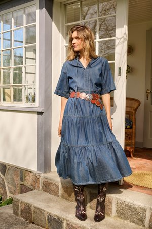 Femme portant une robe en denim bleu avec une large ceinture et des bottes noires brodées, debout sur des marches en pierre devant la porte d'une maison.