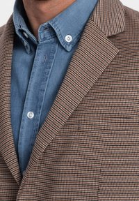 Brązowo-czarna kraciasta marynarka z klapami z wycięciem, zestawiona z jasnoniebieską koszulą zapinaną na guziki. Tkanina wydaje się mieć fakturę, marynarka dopasowana.