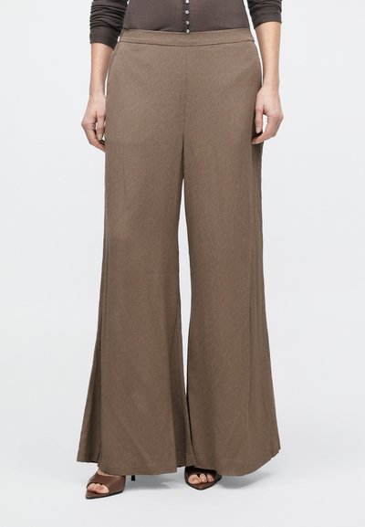 Donna che indossa pantaloni marroni a gamba larga e tacchi aperti marroni, in piedi contro uno sfondo chiaro e uniforme.