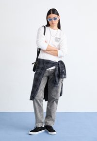 Cămașă albă cu mânecă lungă și logo, blugi gri din denim, cămașă pătrată neagră legată la talie, adidași negri și ochelari de soare bleu deschis.