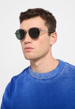 Giovane uomo con capelli castani corti, che indossa occhiali da sole rotondi neri e un maglione blu girocollo, che guarda leggermente di lato.