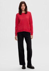 Rode wollen sweater met een ronde halslijn, lange mouwen en een iets verkorte zoom. Gepaard met zwarte wijde jeans en zwarte schoenen.