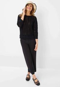 Schwarzer gestrickter Pullover mit offener Masche, dreiviertelärmlig, kombiniert mit schwarzen Hosen mit floralem Stickmuster am Saum und schwarzen Mesh-Schuhen.