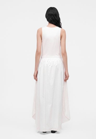 Femme aux longs cheveux noirs portant un haut blanc sans manches et une jupe blanche fluide taille haute, debout dos au spectateur.