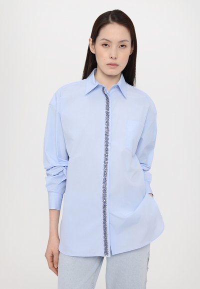 Light blue shirt with long sleeves, a pointed collar, and a decorative patterned trim along the button placket. Features a chest pocket.