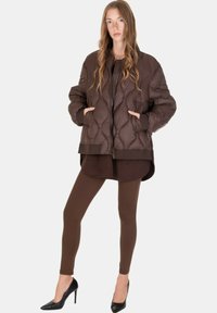 Donna in piedi con le mani nelle tasche, indossa una giacca marrone trapuntata, leggings abbinati e scarpe nere con tacchi alti su uno sfondo semplice.