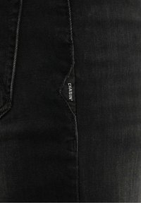 Jeans en denim noir présentant un design cousu, une étiquette latérale avec du texte et une texture lisse.