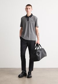 Sivý polo tričko s čiernym golierom, s malým logom. Čierne nohavice a topánky. Drží veľkú čiernu športovú tašku. Minimalistické pozadie.