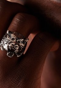 Silver tiger head ring on a hand, featuring detailed facial features and a textured band, set against brown skin.