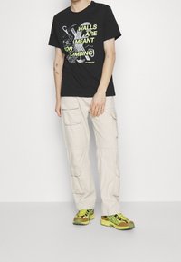 Černé bavlněné tričko s grafikou "ZDI JSOU URČENY K LEZENÍ" v neonově zelené barvě; kombinováno se béžovými cargo kalhotami a vícebarevnými sportovními botami.