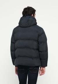Mand iført en sort vinterjakke med hætte, vendt væk, parret med mørkeblå jeans, mod en ensfarvet hvid baggrund.