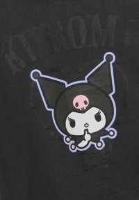 T-shirt noir avec un personnage de dessin animé portant un chapeau noir et un détail de skull rose. Le design comprend un geste de "chut" et des contours audacieux.