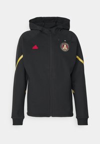 Svart hoodiejacka med gula accenter och en broderad Atlanta United FC-logotyp. Tillverkad av ett mjukt, flexibelt material med full dragkedja.