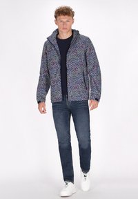 Jachetă ușoară, multicoloră, cu model și glugă, purtată peste o cămașă bleumarin, asortată cu blugi închis și adidași albi.
