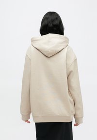 Μπεζ φαρδιά φούτερ με κουκούλα και κορδόνι, μακριά μανίκια και ριμπ μανσέτες. Μαλακό ύφασμα με ομαλή υφή. Μαύρο κάτω ένδυμα.