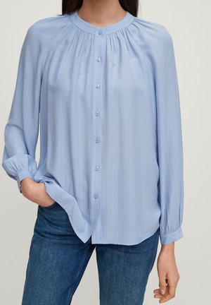 Blouse bleu clair à manches longues avec col rond et boutons sur le devant, portée avec un jean bleu, une main dans la poche.