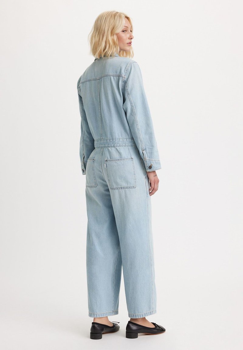 Lyseblå denim-jumpsuit med lange ærmer, knaplukning ved manchetter, elastisk talje, to baglommer, vidbenet design og ensartet tekstur.
