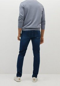 Men's dark blue denim jeans with a slim fit, worn with a light gray long-sleeve shirt. Shoes are light with a beige accent.