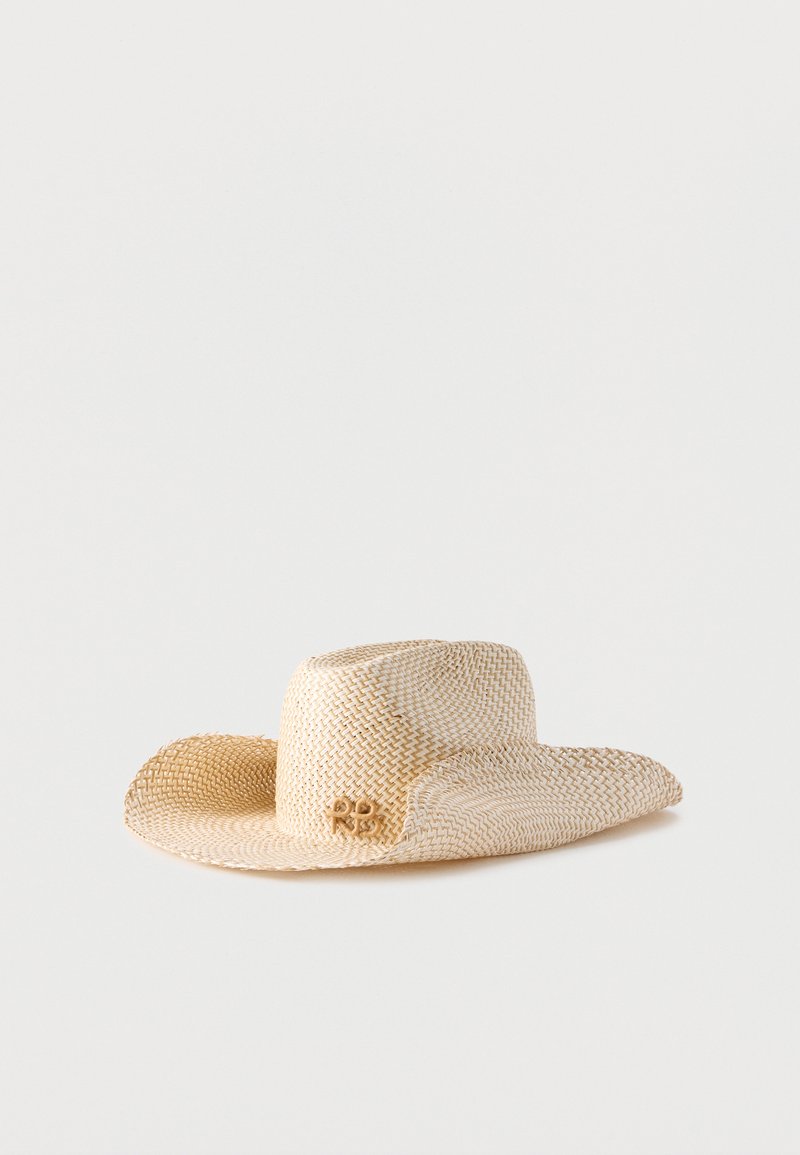 Ανοιχτό μπεζ πλεκτό καπέλο από ψάθα με πλατύ γείσο ελαφρώς καμπυλωτό και μικρό διακοσμητικό έμβλημα στη μία πλευρά.
