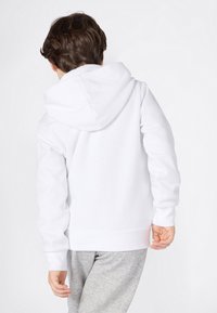Barn med brunt hår iført en hvit hettegenser og grå joggebukser, står med ryggen til og den ene hånden litt krummet.