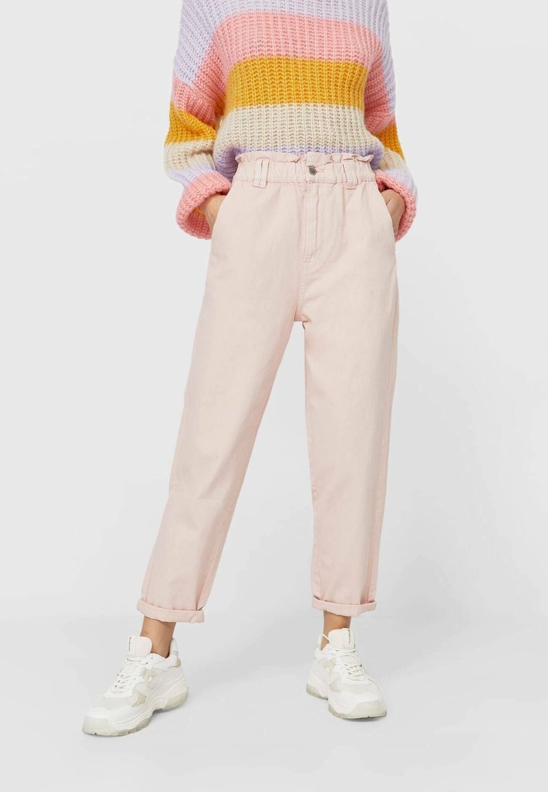 Personne portant un pantalon rose clair taille haute avec revers roulés, un pull en maille rayé pastel et des baskets blanches épaisses, debout les mains dans les poches.