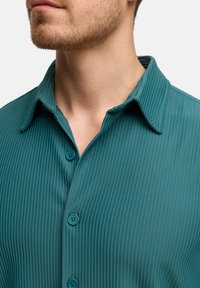 T-Shirts in Petrol mit einem Hemdkragen, aus strukturiertem Stoff und zwei passenden Knöpfen an der Frontleiste.