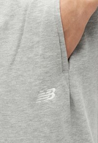 Χέρι μερικώς μέσα στην πλαϊνή τσέπη γκρι αν οιχτόχρωμου φερμουάρ φόρμας με λευκό λογότυπο New Balance κεντημένο κοντά στην τσέπη.