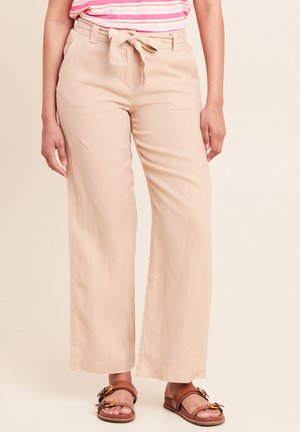 Vrouw draagt beige, hooggetailleerde wijde broek met een stoffen riem voorgebonden en bruine platte sandalen met dubbele gesp.
