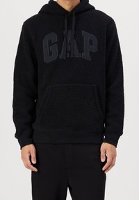 Person mit einer strukturierten schwarzen Kapuzenjacke mit Kängurutasche und erhöhten "GAP"-Buchstaben auf der Brust, kombiniert mit schwarzen Hosen.