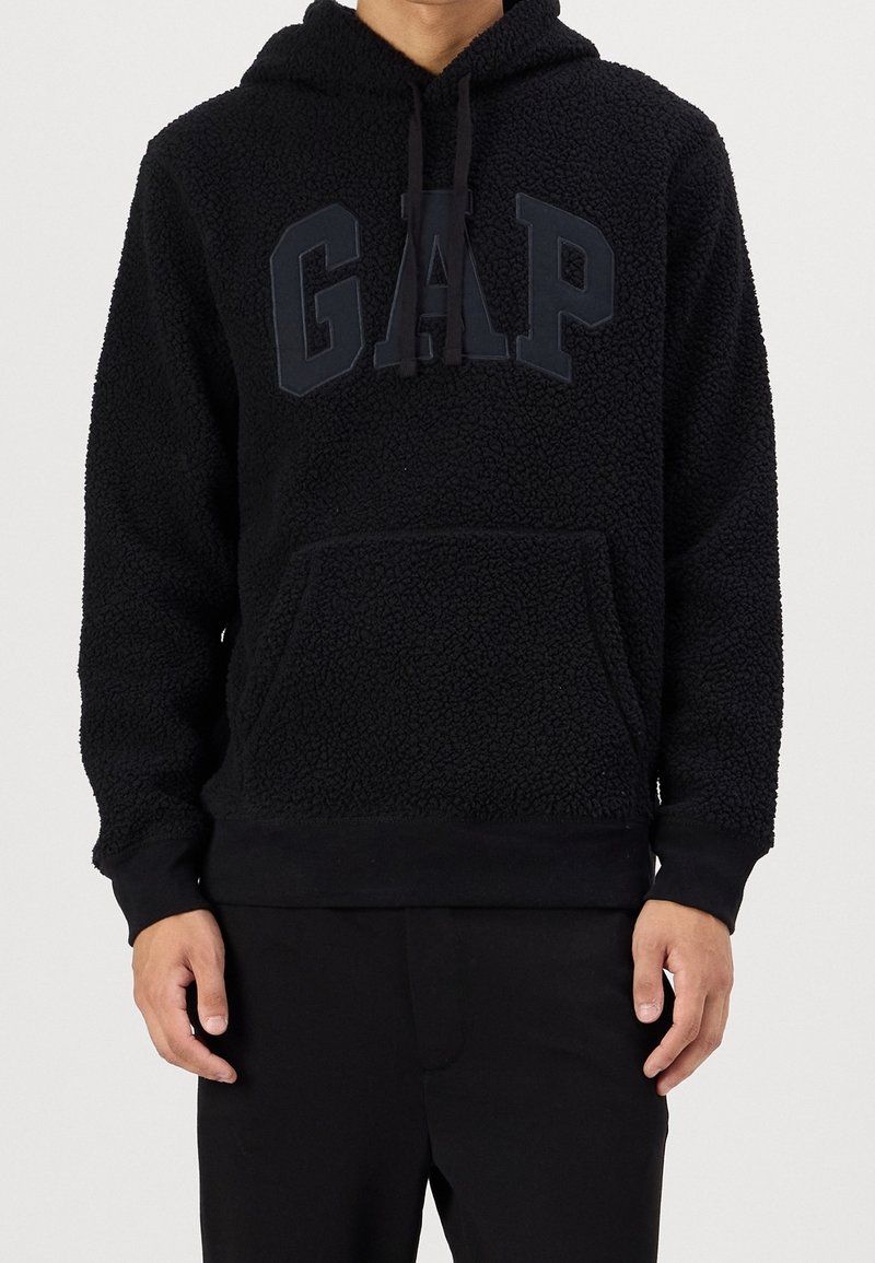 Person mit einer strukturierten schwarzen Kapuzenjacke mit Kängurutasche und erhöhten "GAP"-Buchstaben auf der Brust, kombiniert mit schwarzen Hosen.